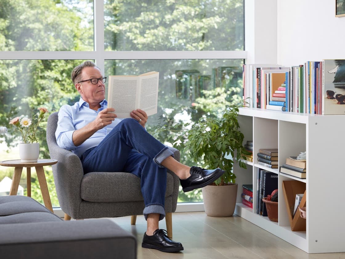 A man reading in an armchair beside a sunlit window and a bookcase