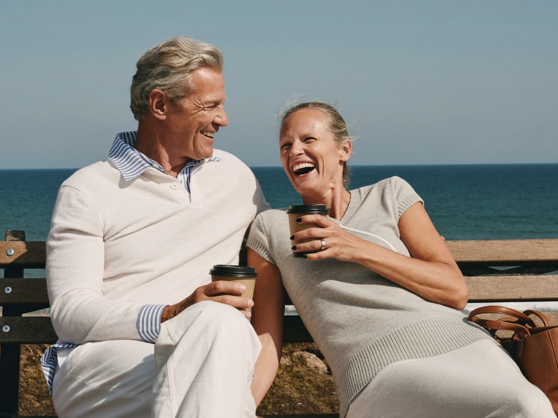 A retired couple laughing together on a bench by the sea with takeaway coffees